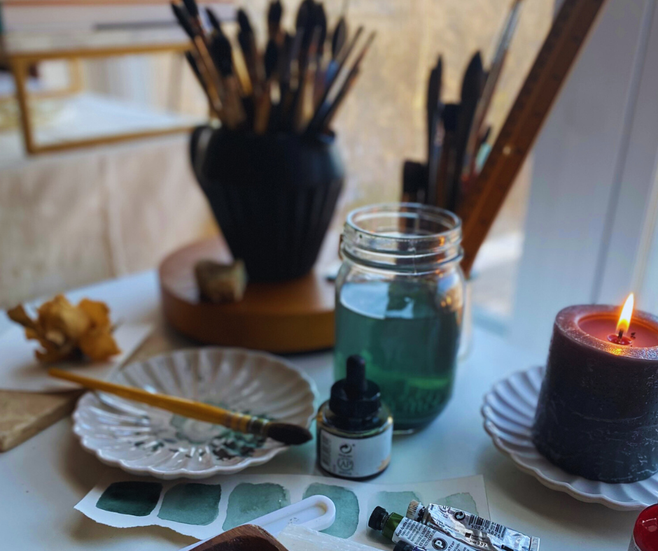 artist table in front of window with paintbrushes a paint palette and a few paints on the table. There is also a candle burning on the table