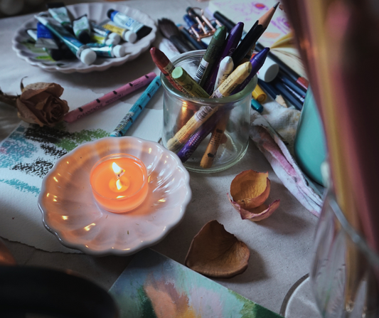 artist table with oil pastels paint tubes makers on the table along with paper a small tea candle and dried flowers