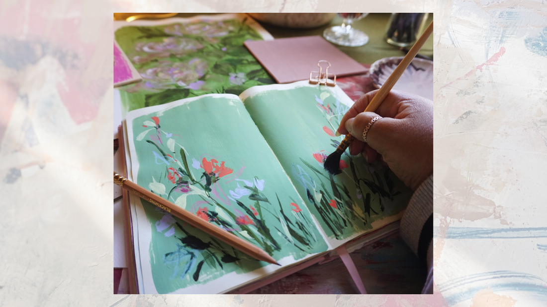 artist desk with artist hand holding a paintbrush while painting in their sketchbook 